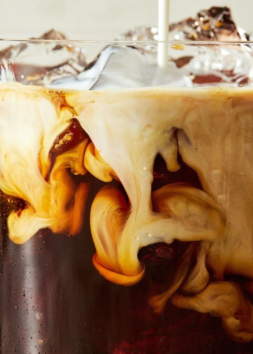 Cream swirling through cold brew coffee over ice, seen close up through the glass