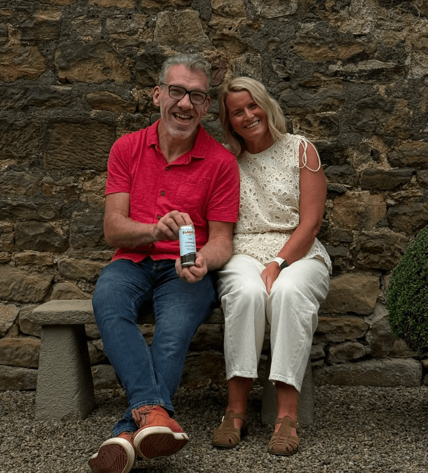 David and Tricia sitting on a stone bench against a drystone wall, holding a can of Iced Caramel Cold Brew